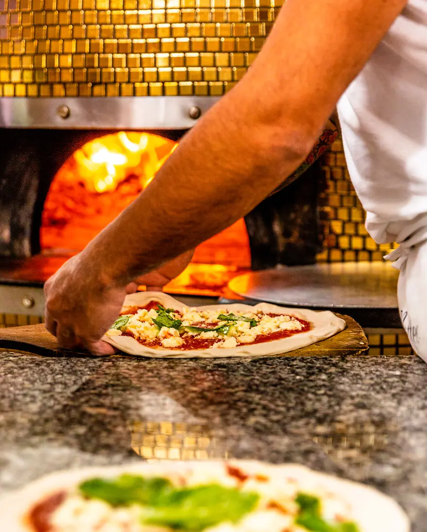 EIn Hand zieht eine fertig belegte Pizza auf den Pizzaschieber, im Hintergrund brennt ein goldverzierter Kuppelofen.